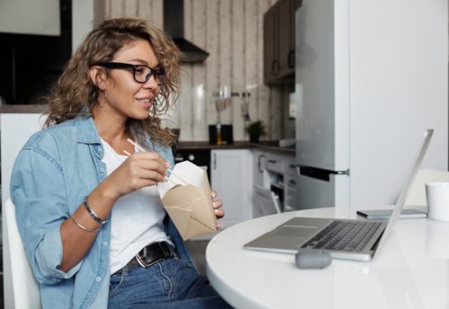 woman eating takeout food and looking at laptop