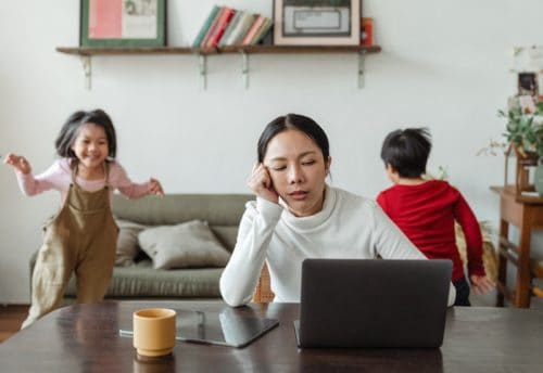 Fatigued woman on laptop with running kids behind her